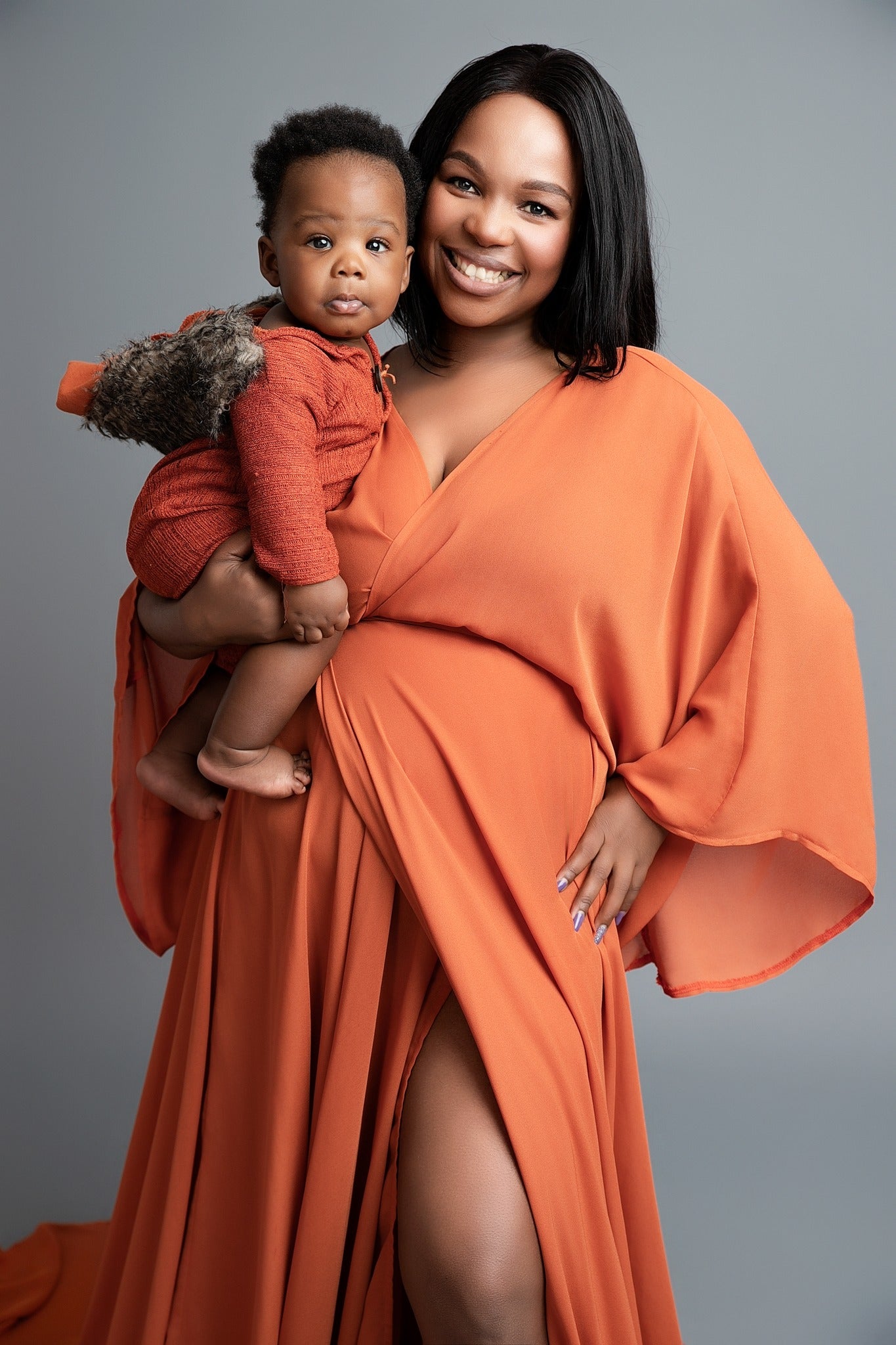 A woman in a orange dress holding a baby who is wearing a brown outfit. They are indoors with a gray background.