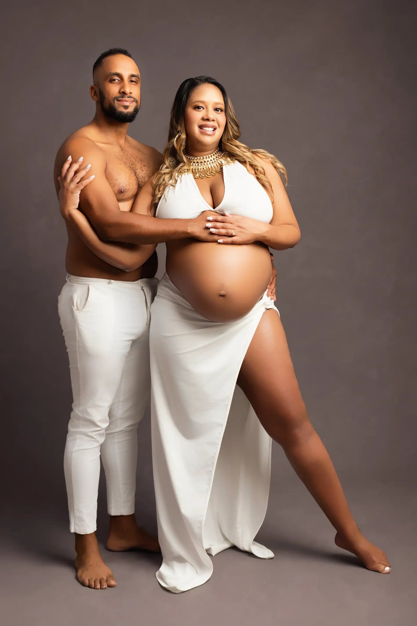 A pregnant woman and her partner posing for a maternity photo shoot, both dressed in white, with the woman in the foreground holding her belly.