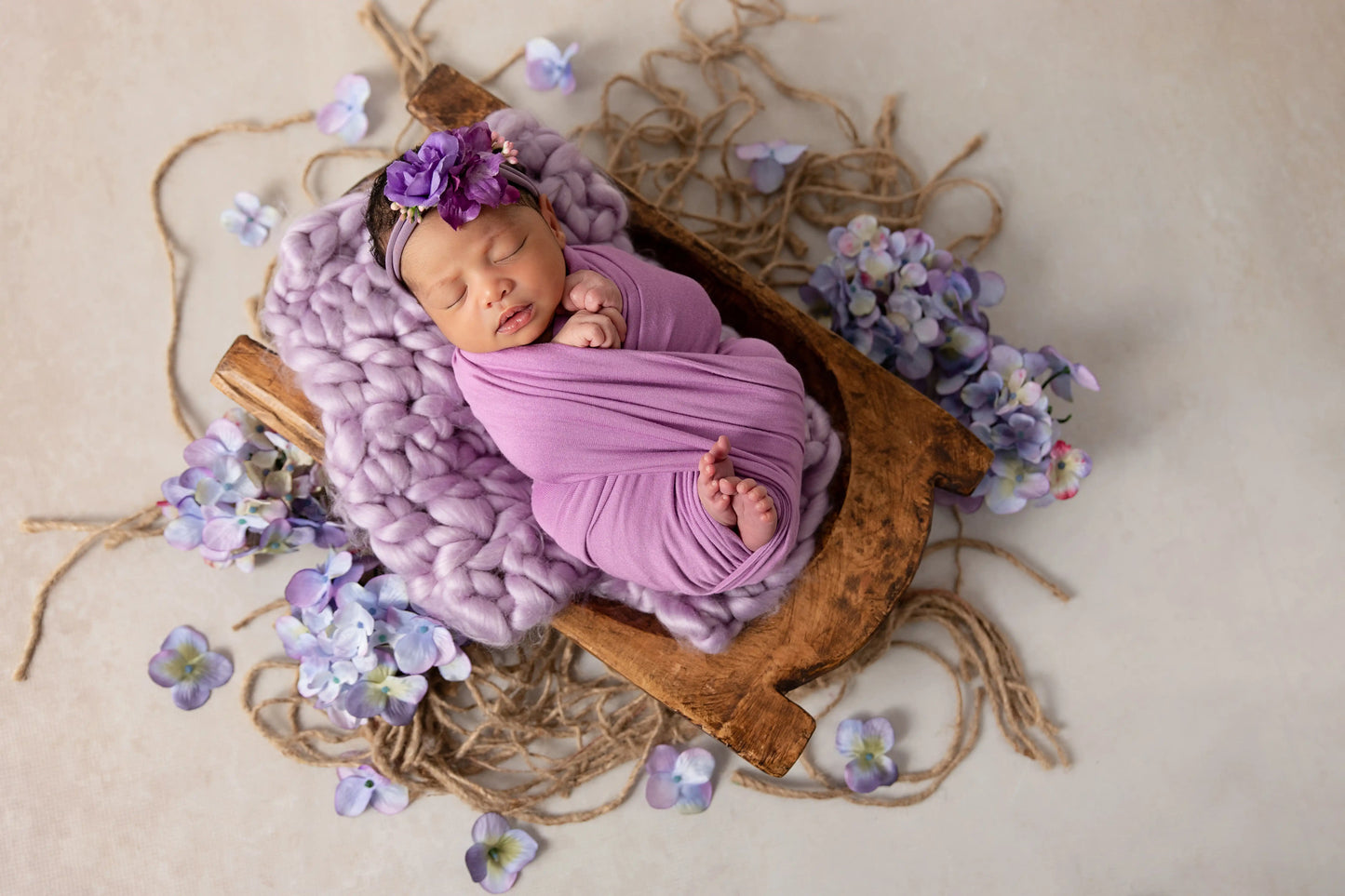 Newborn baby wrapped in purple fabric lying in a wooden basket with flowers around.