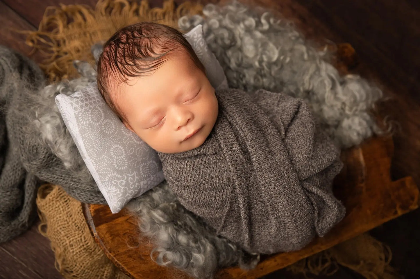Newborn baby wrapped in gray fabric on a wooden surface with textured blankets