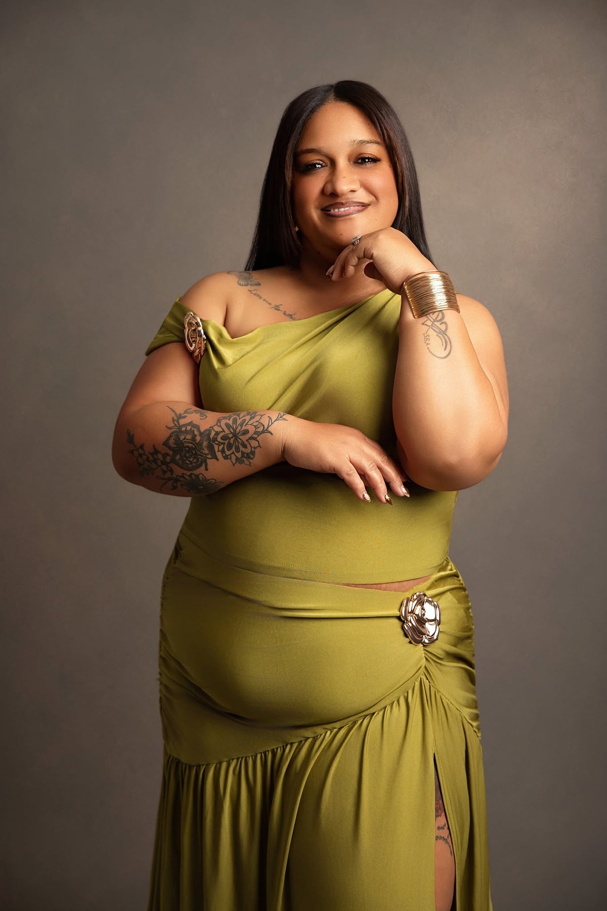 A woman posing in a draped olive green dress with a large decorative brooch at the waist, set against a solid dark gray studio background.
