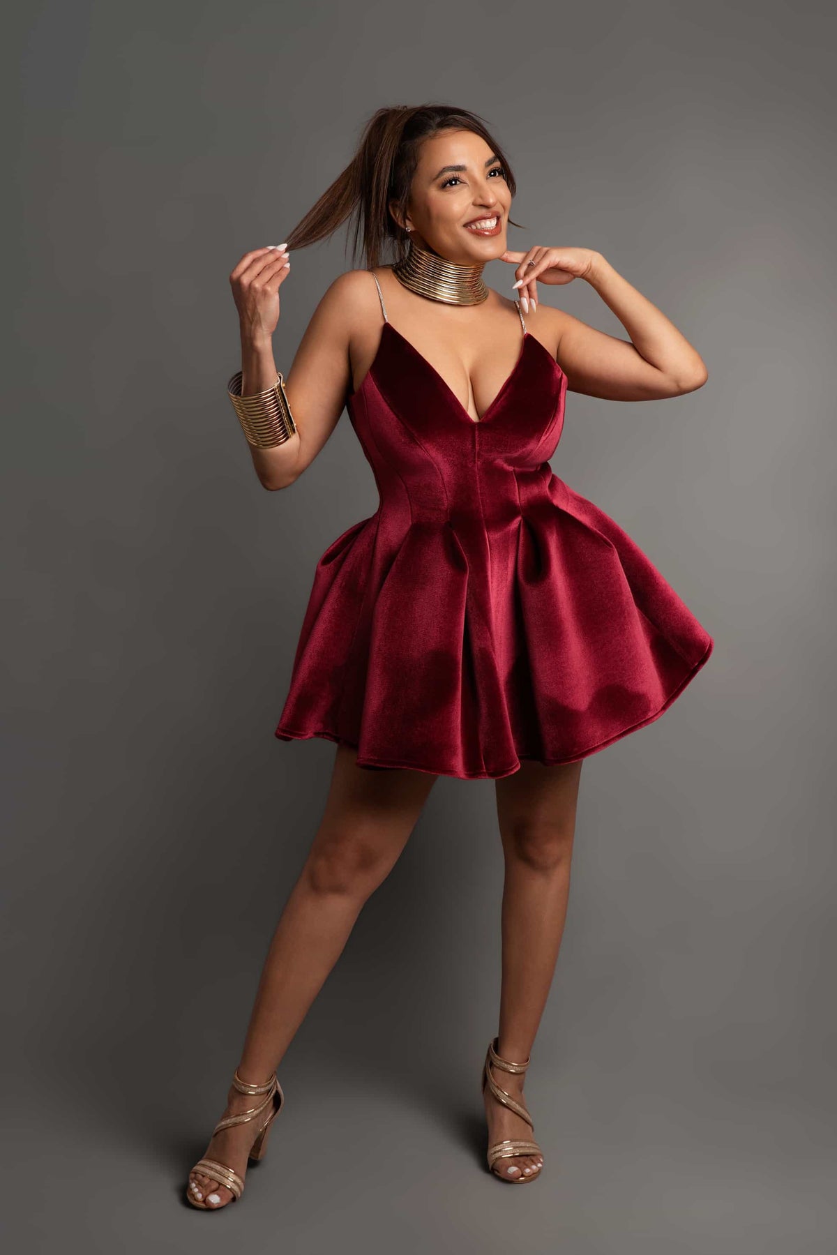 A woman modeling a short, deep burgundy velvet mini dress with spaghetti straps, a plunging neckline, and a flared skirt, styled with metallic gold accessories against a solid gray studio background.