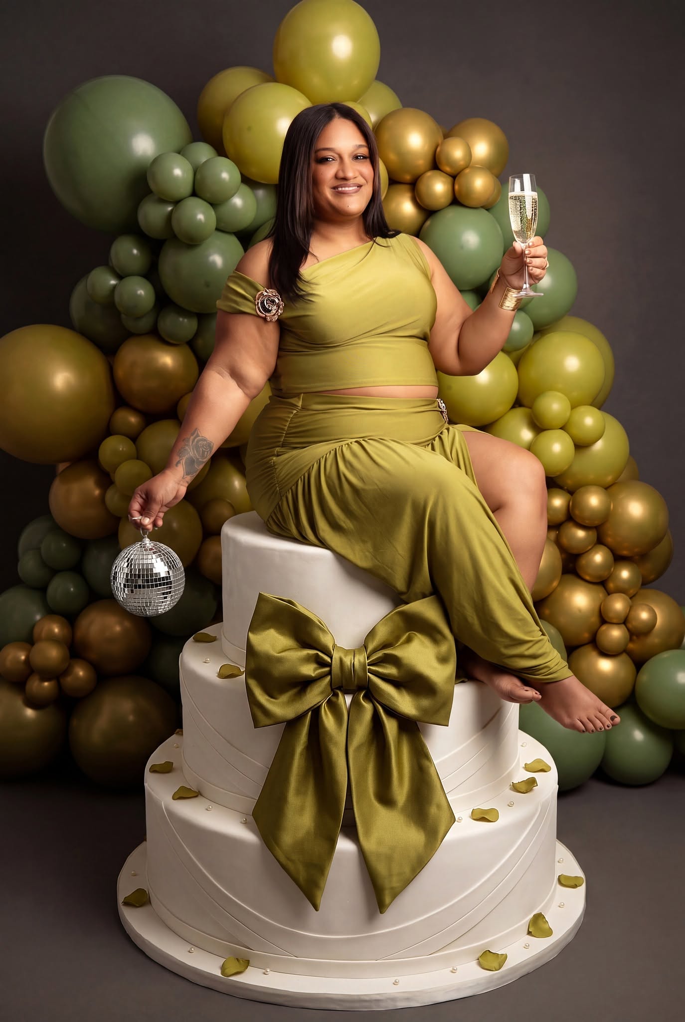 A woman wearing an olive green two-piece outfit sits on a white tiered cake decorated with a large matching green bow, holding a champagne flute and a disco ball against a backdrop of green and gold balloons.