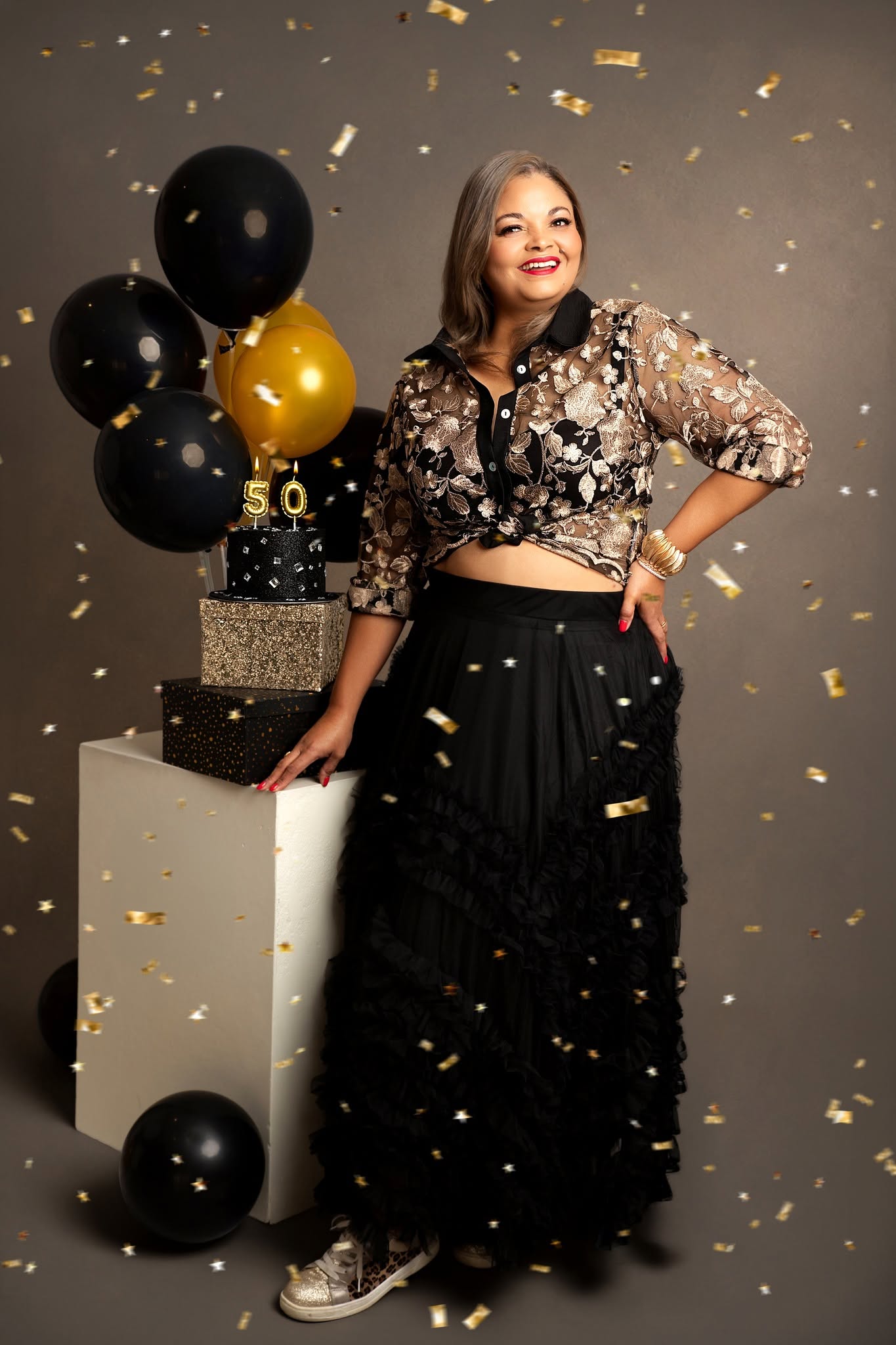 A woman posing in a black and gold sequined crop top and a long black ruffled skirt, standing next to a white pedestal decorated with black and gold balloons and gold confetti against a dark gray background.