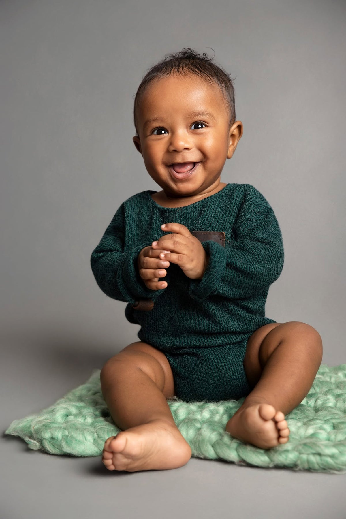 A smiling baby sitting on a textured mint green blanket against a solid gray studio background, wearing a dark teal-green knit long-sleeve romper.