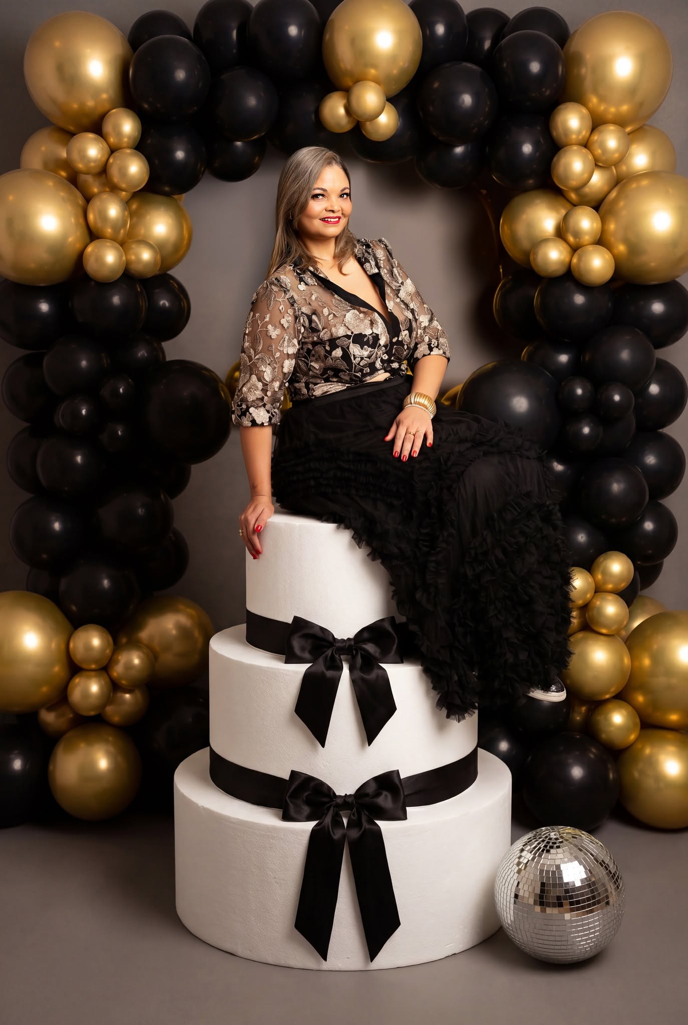 A woman with gray hair sits on a three-tier white cake decorated with black ribbons and bows, posed in front of a black and gold balloon arch with a silver disco ball on the floor.