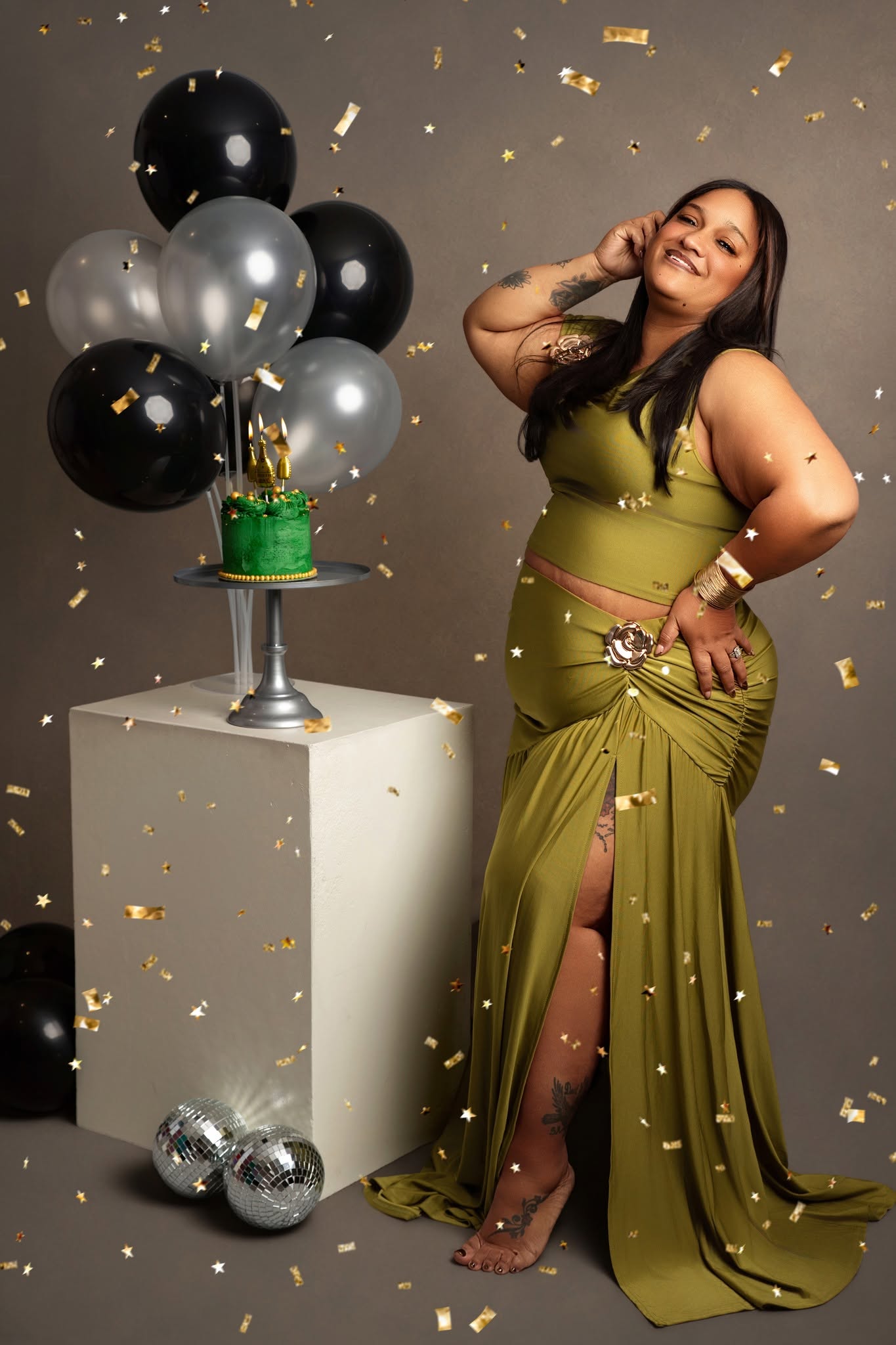 A woman in a flowing olive green two-piece dress poses beside a white pedestal with black and silver balloons, a small green cake, and gold confetti against a taupe studio background.