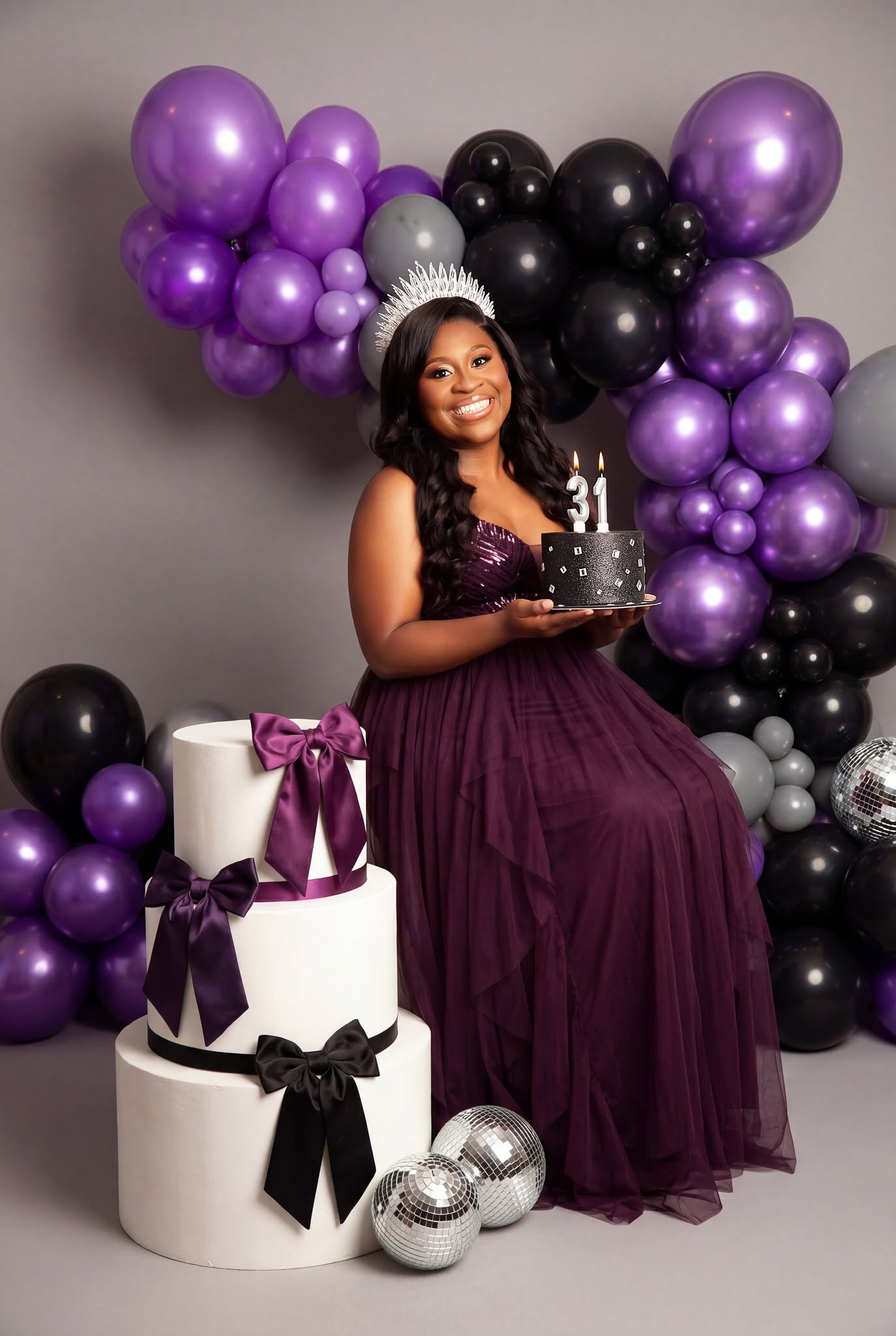 A woman in a purple strapless gown and tiara stands behind a small birthday cake with candles, posed in front of a large balloon arch of purple, black, and silver balloons. A white tiered cake with purple and black ribbons sits in the foreground on a gray studio background.