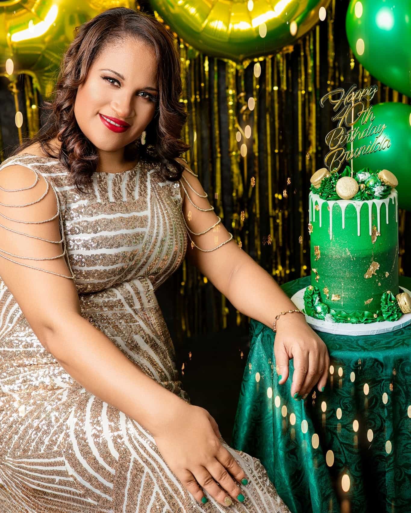 Woman in a sparkly dress posing next to a green cake with gold decorations against a gold and green background.