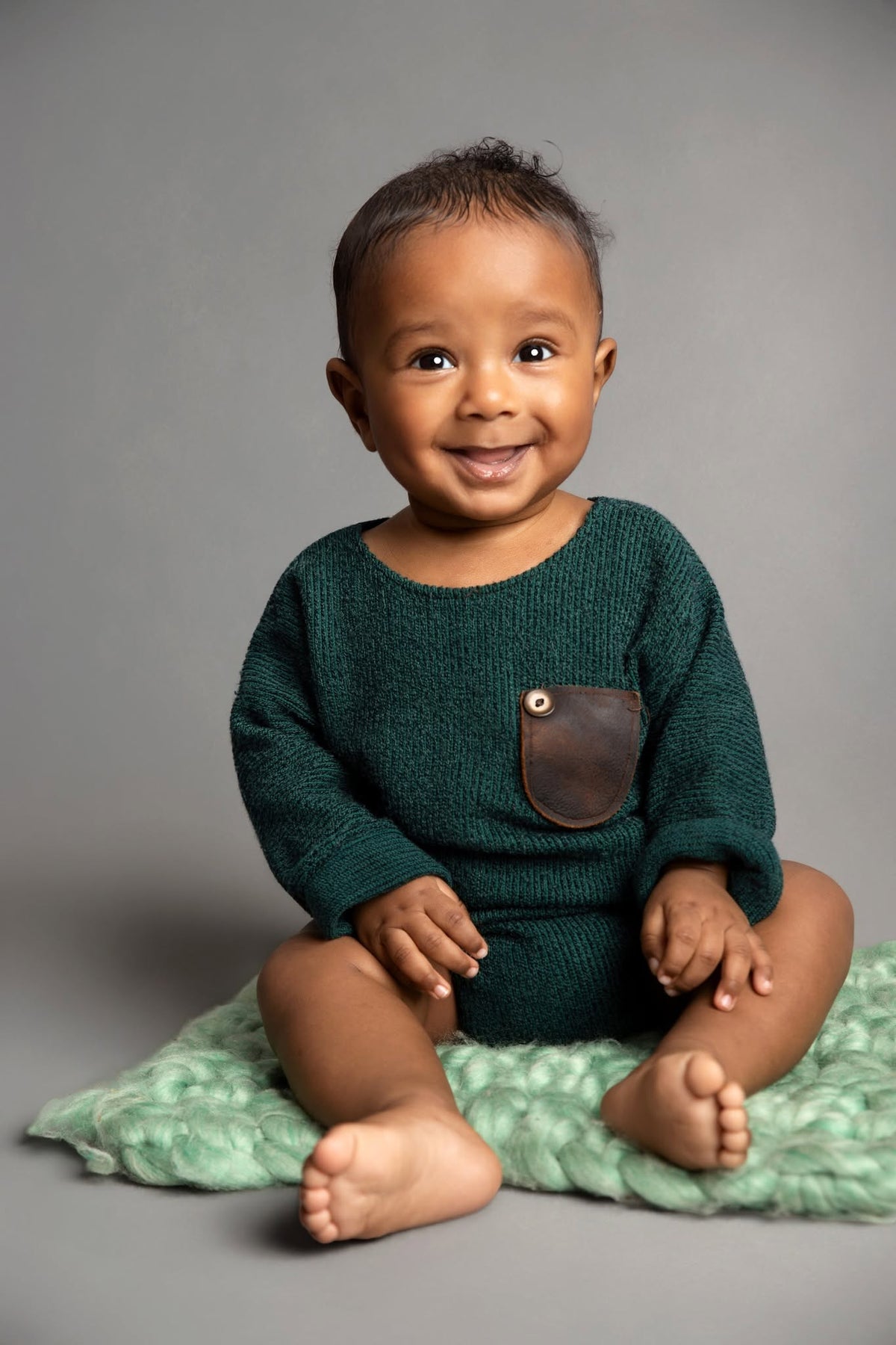 A smiling baby sitting on a textured mint green blanket against a solid gray studio background, wearing a dark green knit romper with a brown leather chest pocket.