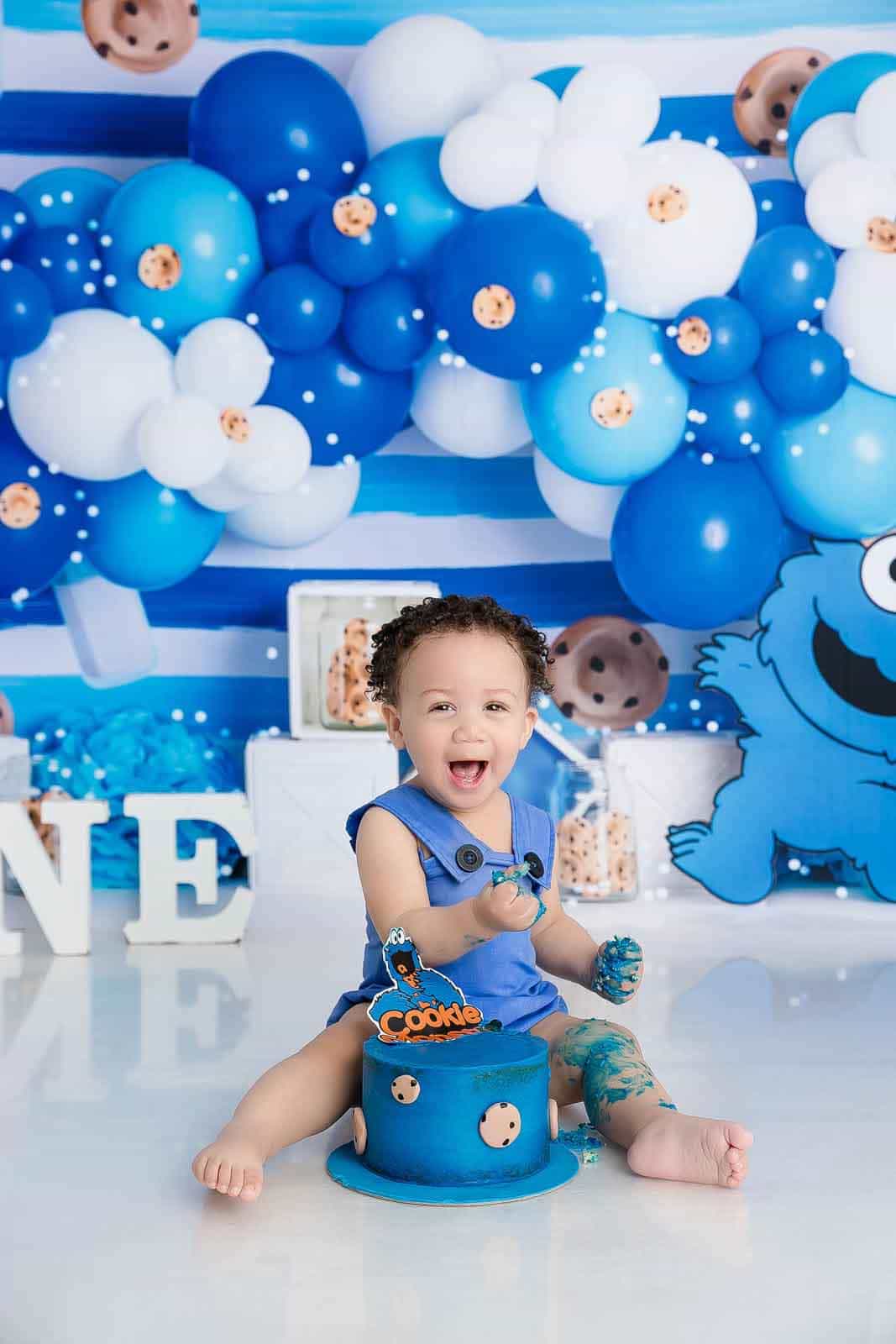 A toddler wearing a blue Cookie Monster-themed outfit sits on a blue cake stand in front of a blue and white balloon backdrop with a large Cookie Monster character cutout and a decorated blue cake.
