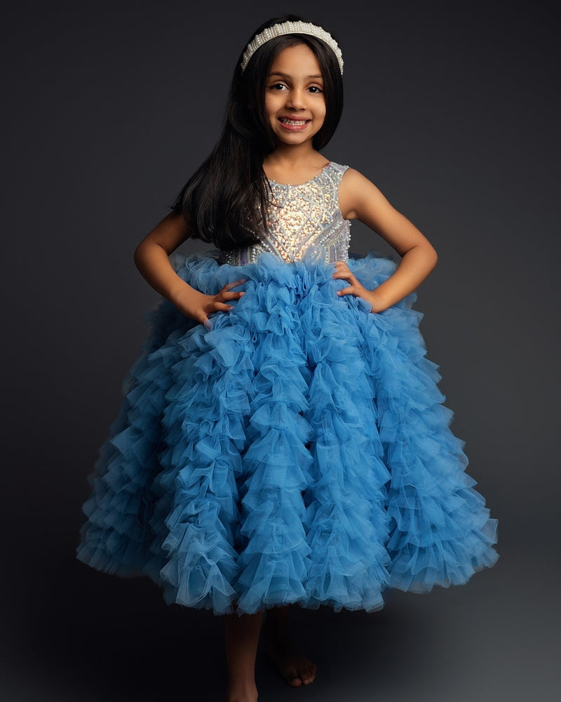A professionally taken photo of a child in a blue tiered dress, posing with hands on hips, smiling at the camera.