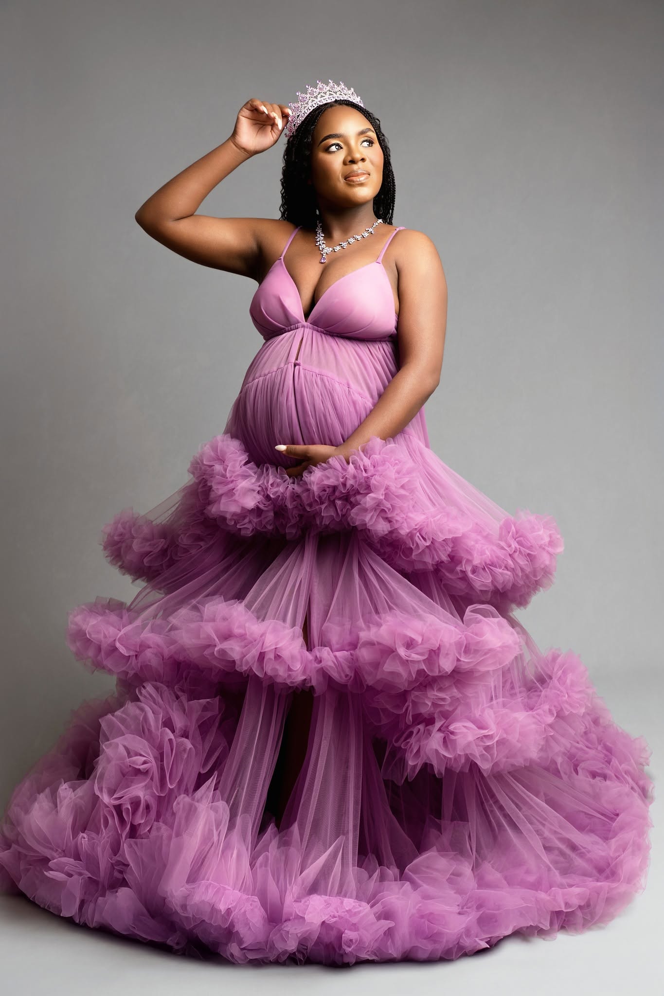 A pregnant woman wearing a voluminous lavender-purple maternity ball gown with layered ruffles and a sweetheart neckline, styled with a tiara and necklace against a solid gray studio background.