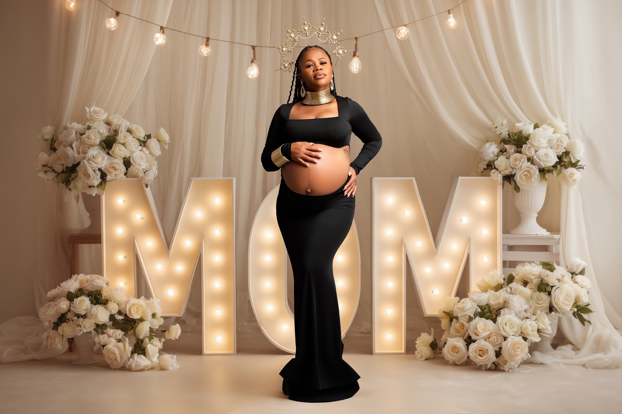 Pregnant woman in a black dress standing in front of 'MOM' letters with floral decorations.