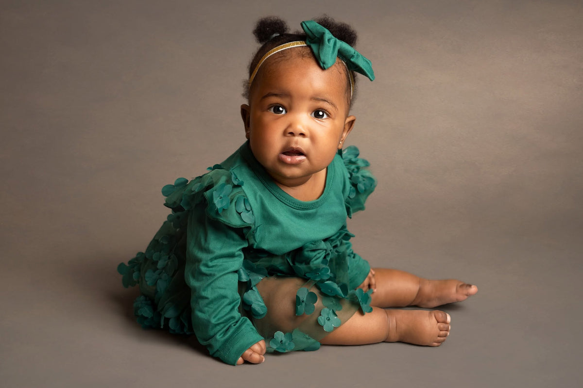 A baby girl sitting on a neutral gray studio background, wearing a textured teal green long-sleeve dress with ruffled details and a matching bow headband.
