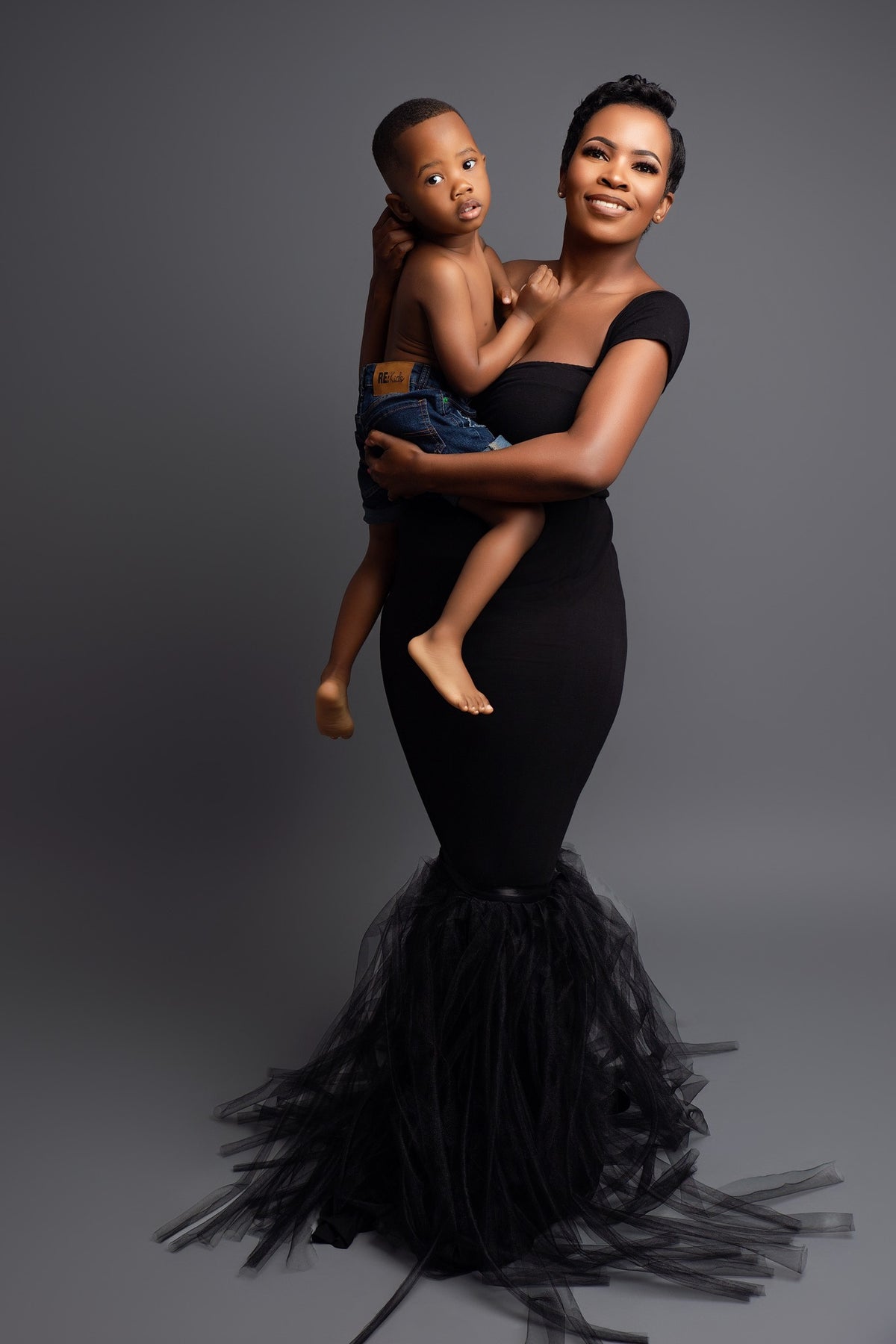 A studio photograph of a mother in a black dress holding her child, posing for a mommy and me photo shoot.