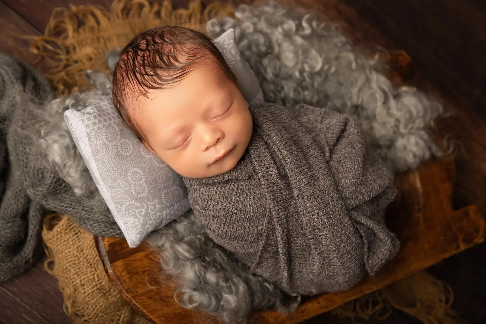 Newborn baby wrapped in gray fabric on a wooden surface with textured blankets