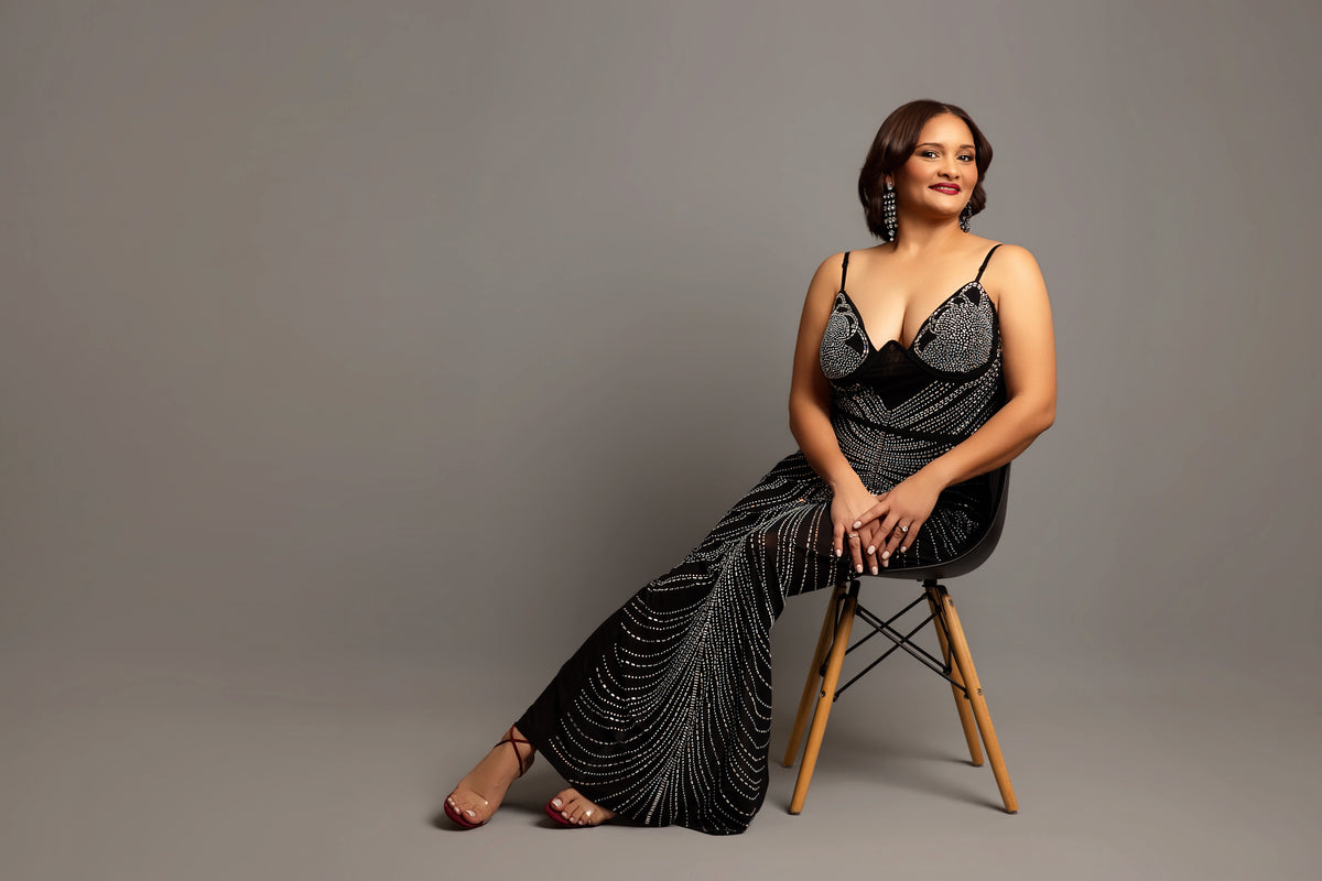 Woman in a black beaded dress sitting on a wooden chair against a gray background