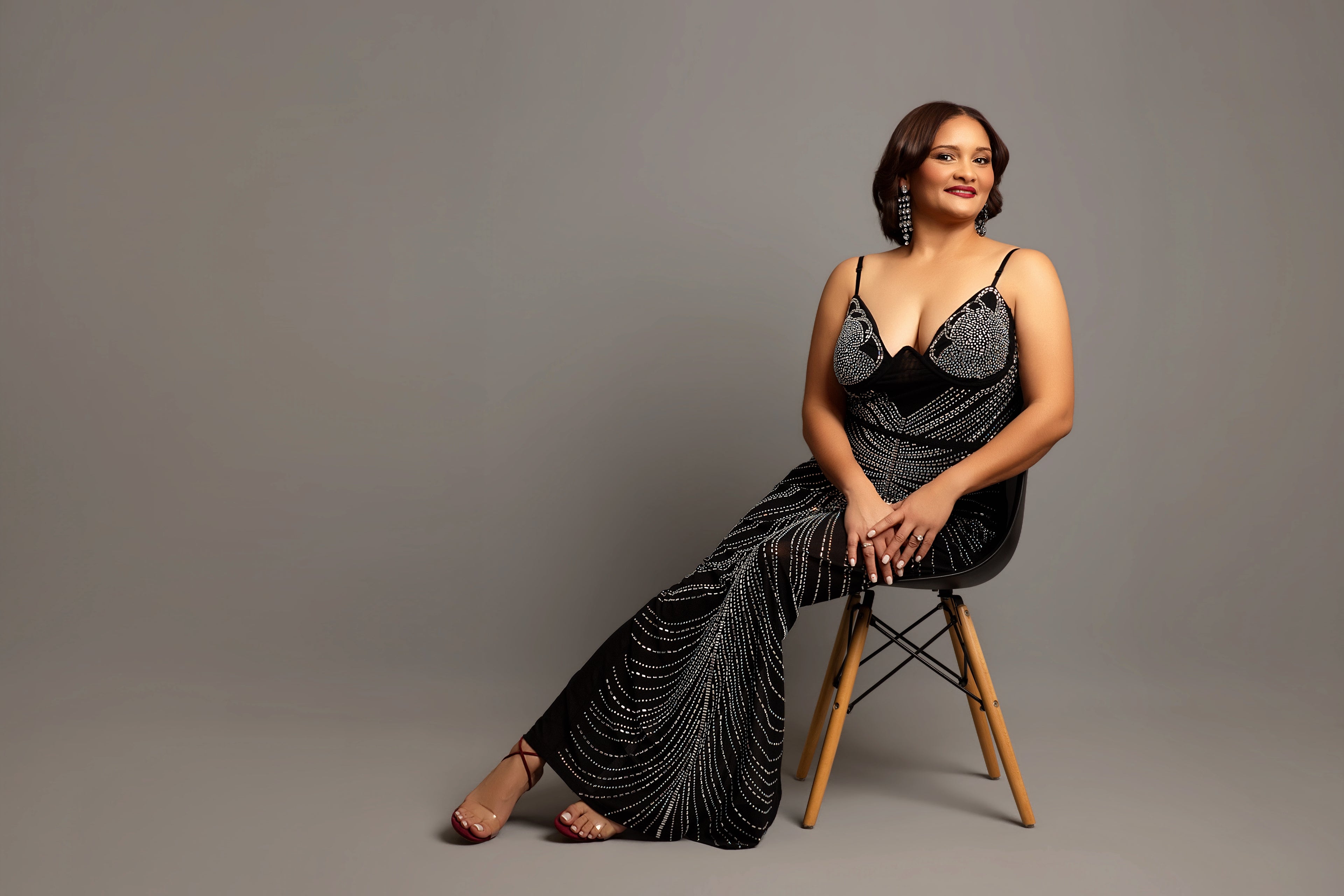 Woman in a black beaded dress sitting on a wooden chair against a gray background