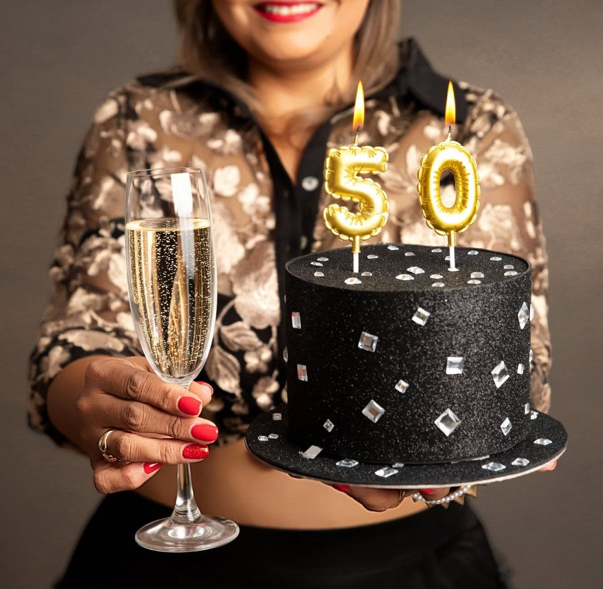 A woman in a patterned blouse holds a black glitter top-hat-shaped cake with silver rhinestone accents and gold candle numbers reading 50, while also holding a filled champagne flute against a plain gray studio background.