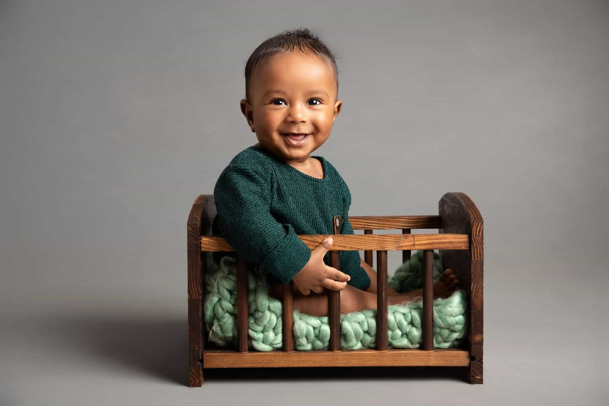 A smiling baby wearing a dark green knit sweater sits inside a small wooden crib with mint green knit bedding, photographed against a plain gray studio background.
