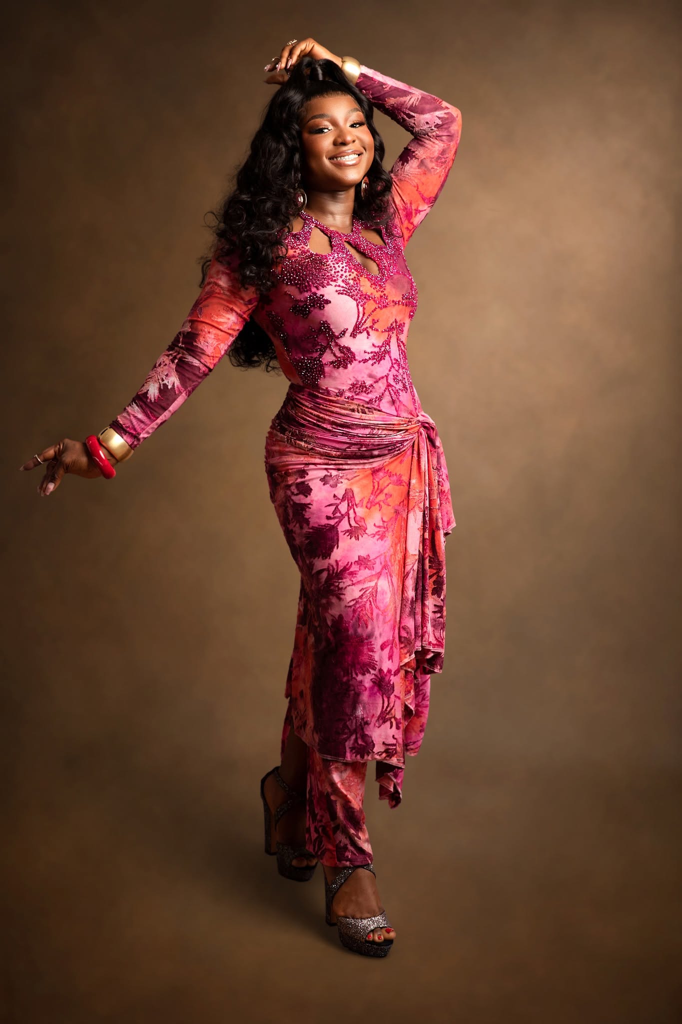 A woman posing in a long-sleeved, knee-length pink and magenta floral dress against a solid brown studio background.