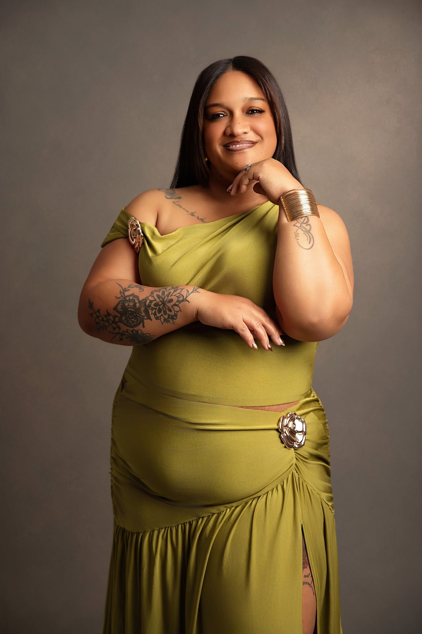 A woman posing in a draped olive green dress with a large decorative brooch at the waist, set against a solid dark gray studio background.