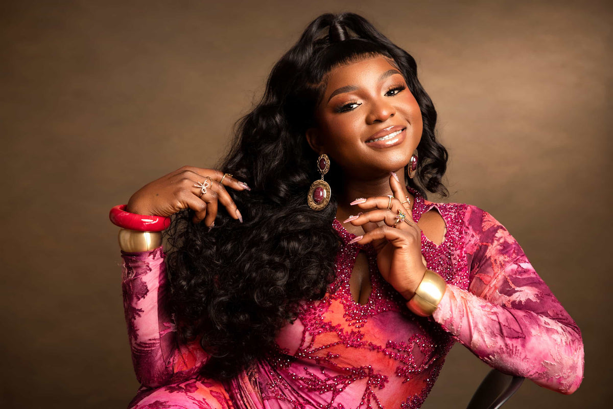 A smiling woman with long, wavy black hair poses in a vibrant pink and magenta patterned dress against a warm brown studio backdrop. She is accessorized with gold jewelry, including a large dangle earring, rings, and bracelets.