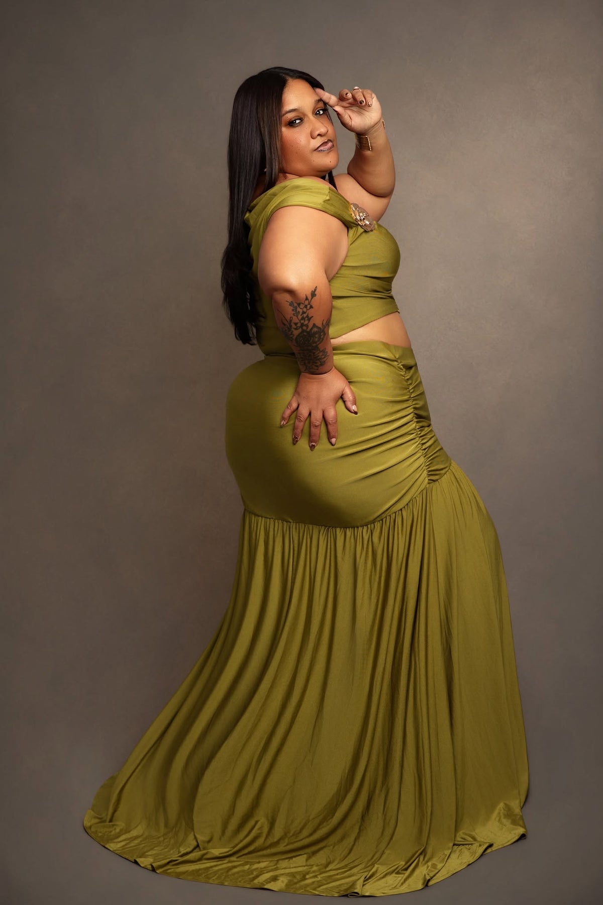 A woman modeling a two-piece olive green dress set against a solid gray studio background. The set features a halter-style crop top with a cutout waist and a matching pleated maxi skirt.
