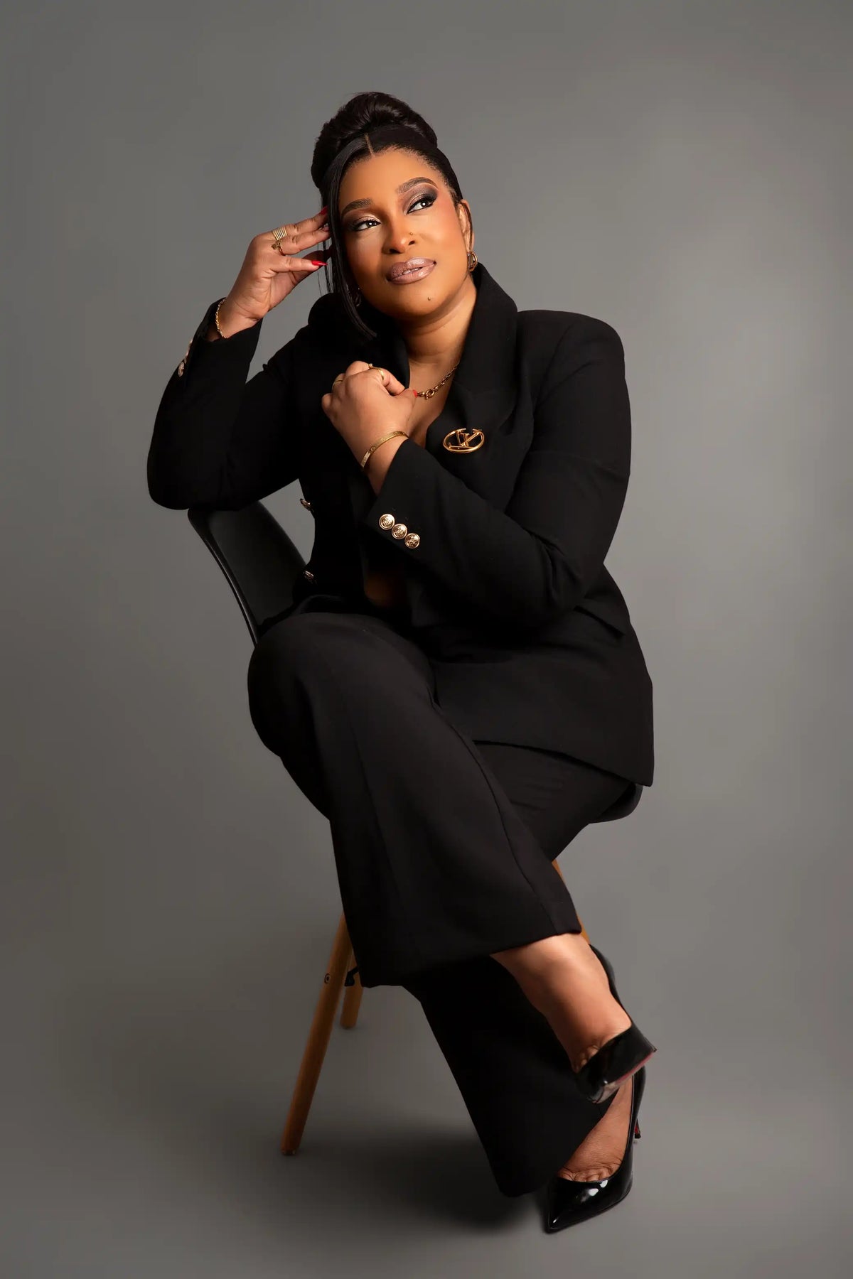 A woman posing in a black pantsuit with a matching blazer, seated on a chair against a solid gray studio background.