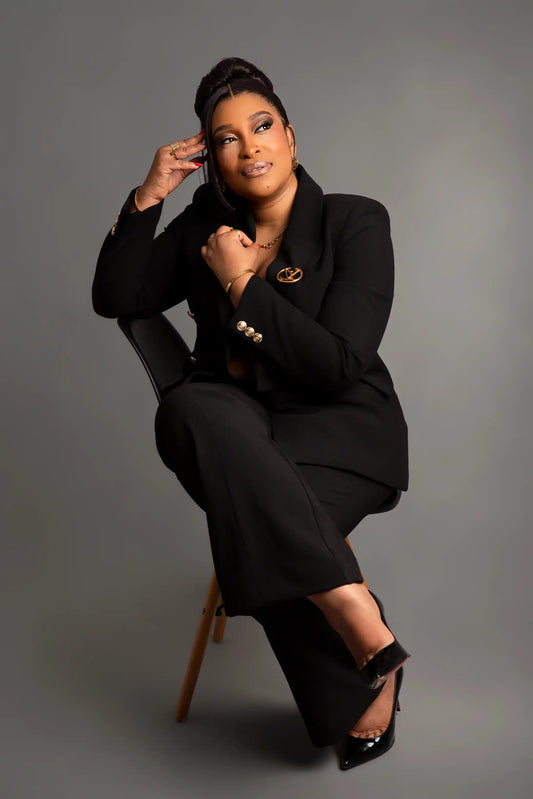 A woman posing in a black pantsuit with a matching blazer, seated on a chair against a solid gray studio background.