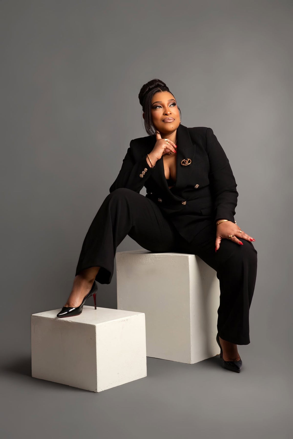 A woman in a black double-breasted blazer and matching trousers sits on white geometric blocks against a solid gray studio background. She is wearing black pointed-toe heels with red soles and has a gold pin on her blazer.